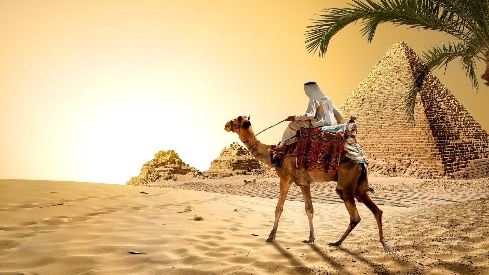 Egy teve hátán ülő ember a sivatagban, a háttérben piramisokkal.