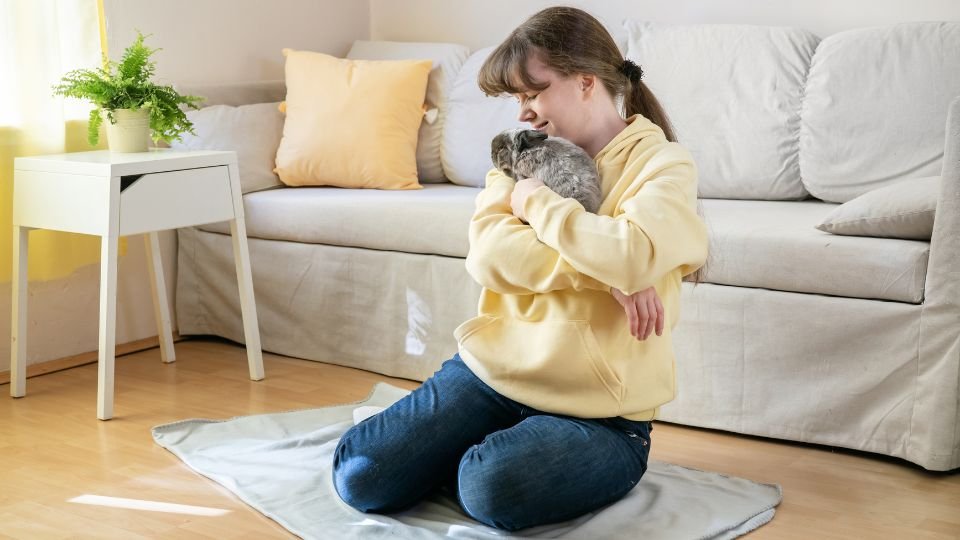 Egy fiatal nő nyulat tart a karjában, miközben a földön ül egy lakásban.