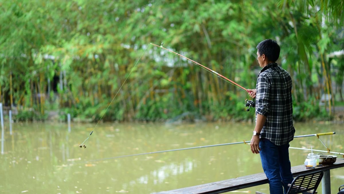 Egy horgász áll a vízparton, horgászbotot tartva a kezében, mögötte zöld növényzet látható.